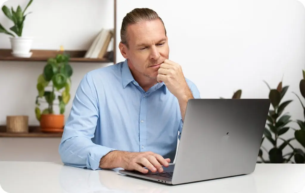 A man in a light blue shirt sits at a desk, looking thoughtfully at a laptop. Shelves with plants and books are visible in the background, creating a modern and tidy workspace.