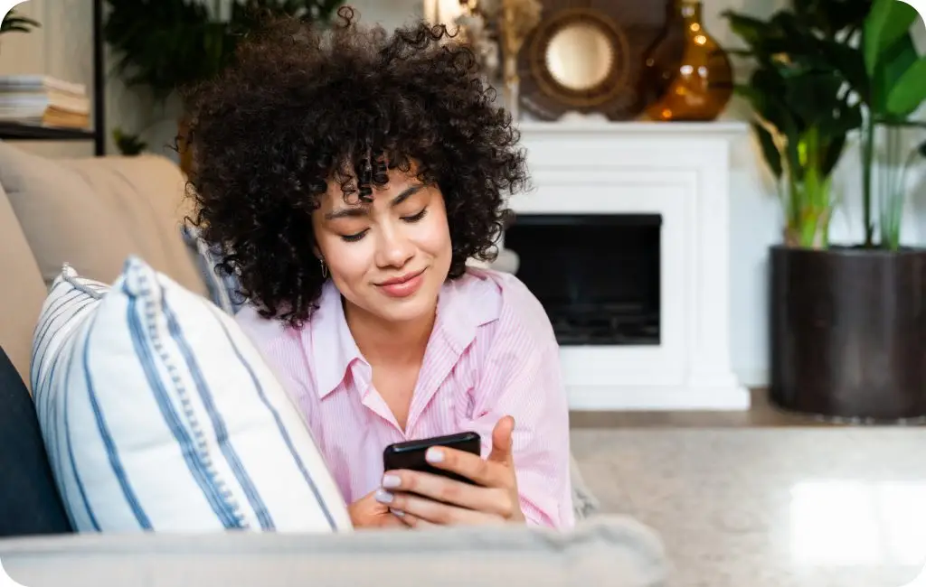 A woman with curly hair wearing a pink shirt sits on a couch, smiling while looking at her smartphone. She is in a cozy living room with plants and decorative items in the background.
