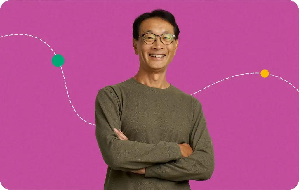A smiling man with glasses and a brown sweater stands with arms crossed in front of a pink background with a dashed white line and colored dots.
