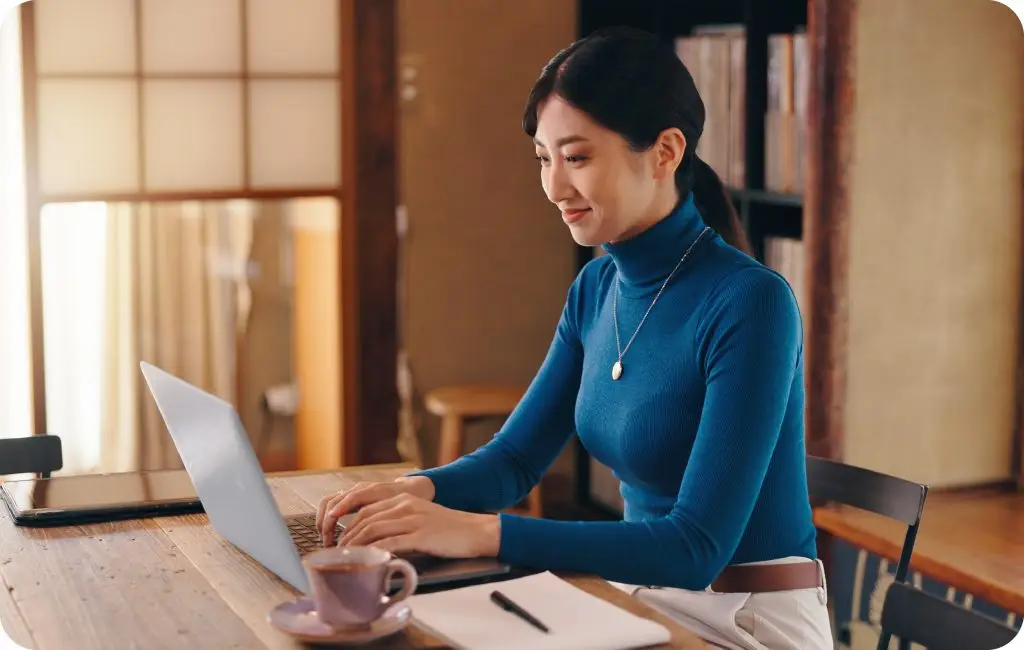 A woman in a blue turtleneck sits at a wooden table, working on a laptop with a notebook and coffee cup beside her, in a cozy room with bookshelves and soft lighting.