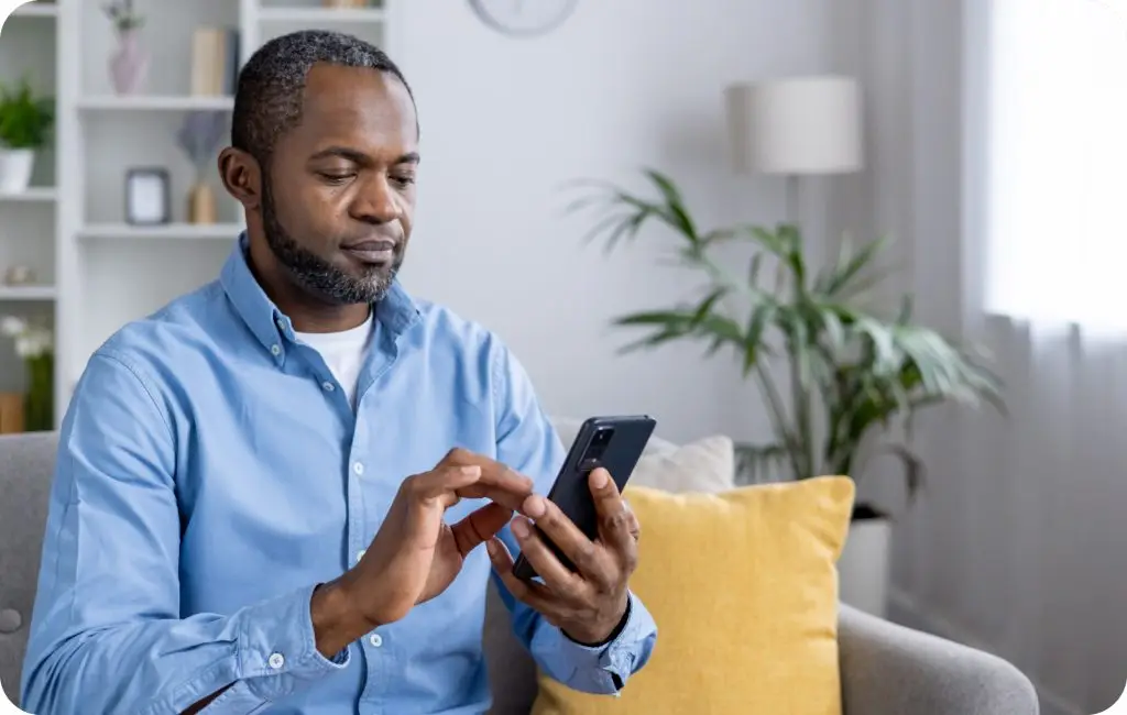 A man sitting on a sofa with a yellow cushion, wearing a light blue shirt, looks at and uses a smartphone in a bright, modern living room with plants and shelves in the background.